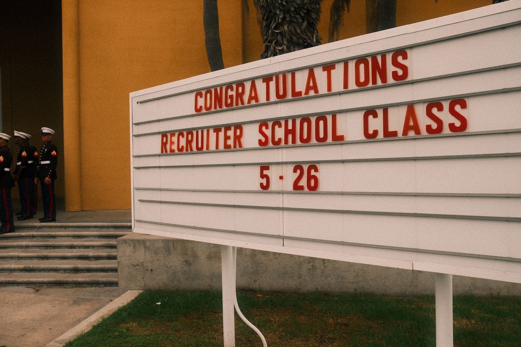 A white letter-board sign reading CONGRATULATIONS RECRUITER SCHOOL CLASS 5-26 in red type, with Marines in dress uniform standing on the steps behind it.