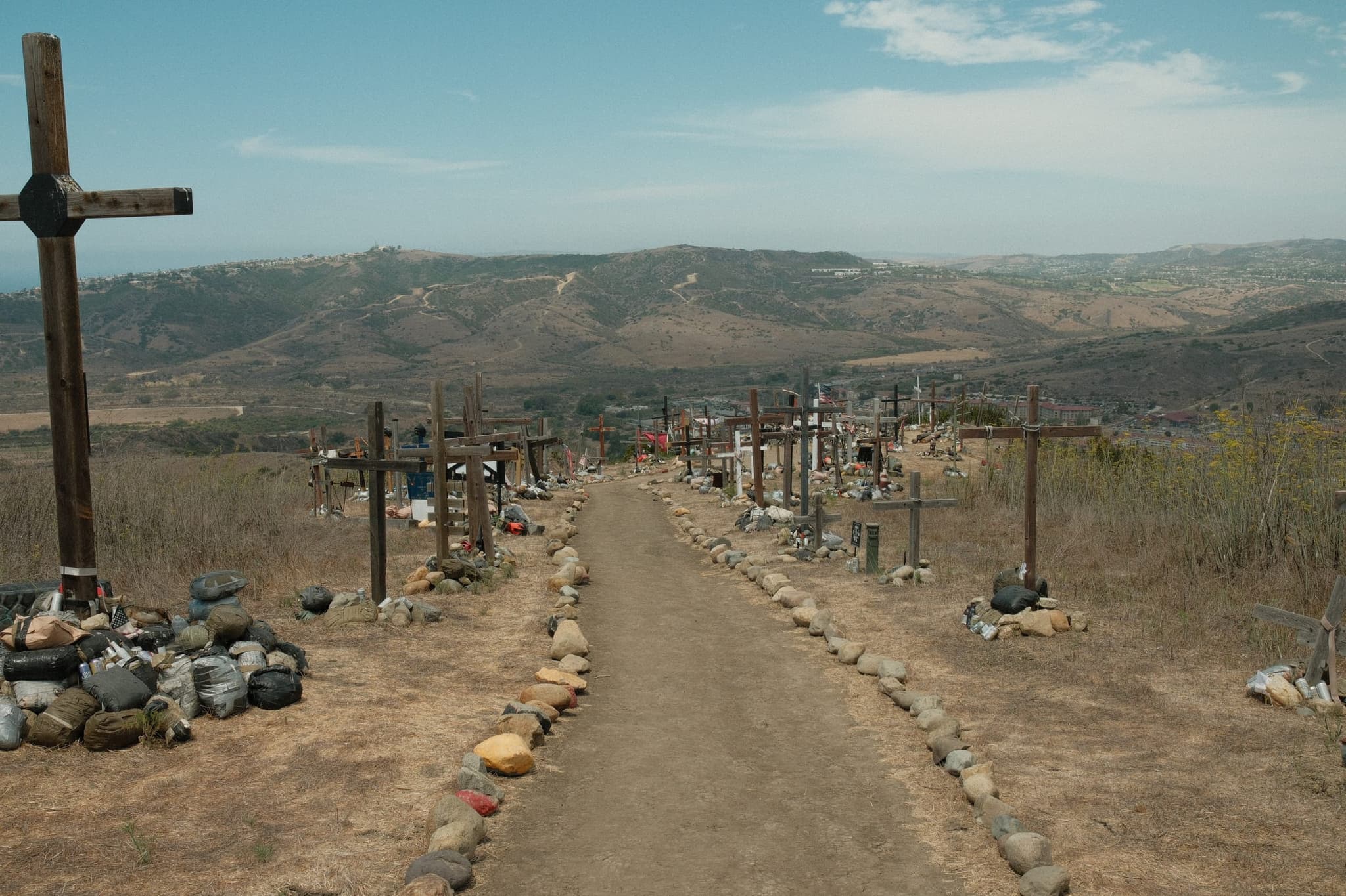 A dirt path running along a dry ridge, edged with rows of wooden crosses and lines of stones, with hills and low cloud beyond.