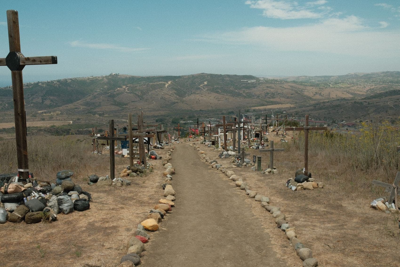 A dirt path running along a dry ridge, edged with rows of wooden crosses and lines of stones, with hills and low cloud beyond.